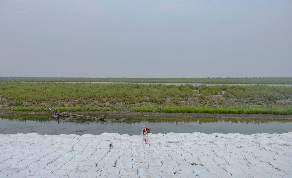 Frauen, die nach dem Sammeln von Wasser zurückkehren und über große Ablagerungen von Geotextiltaschen gehen, die zusammengefügt wurden, um den Damm vor dem ansteigenden Brahmaputra-Fluss vor Bodenerosion während der Hauptmonsunzeit in Indien zu schützen.